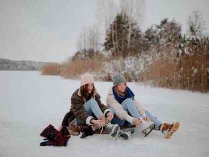 Kaksi nuorta naista laittamassa luistimia jalkaan jäällä. Talvimaisema taustalla.